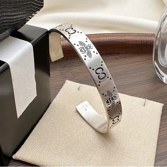 A sterling silver cuff bracelet with a retro bee design, displayed next to its packaging and a perfume bottle.