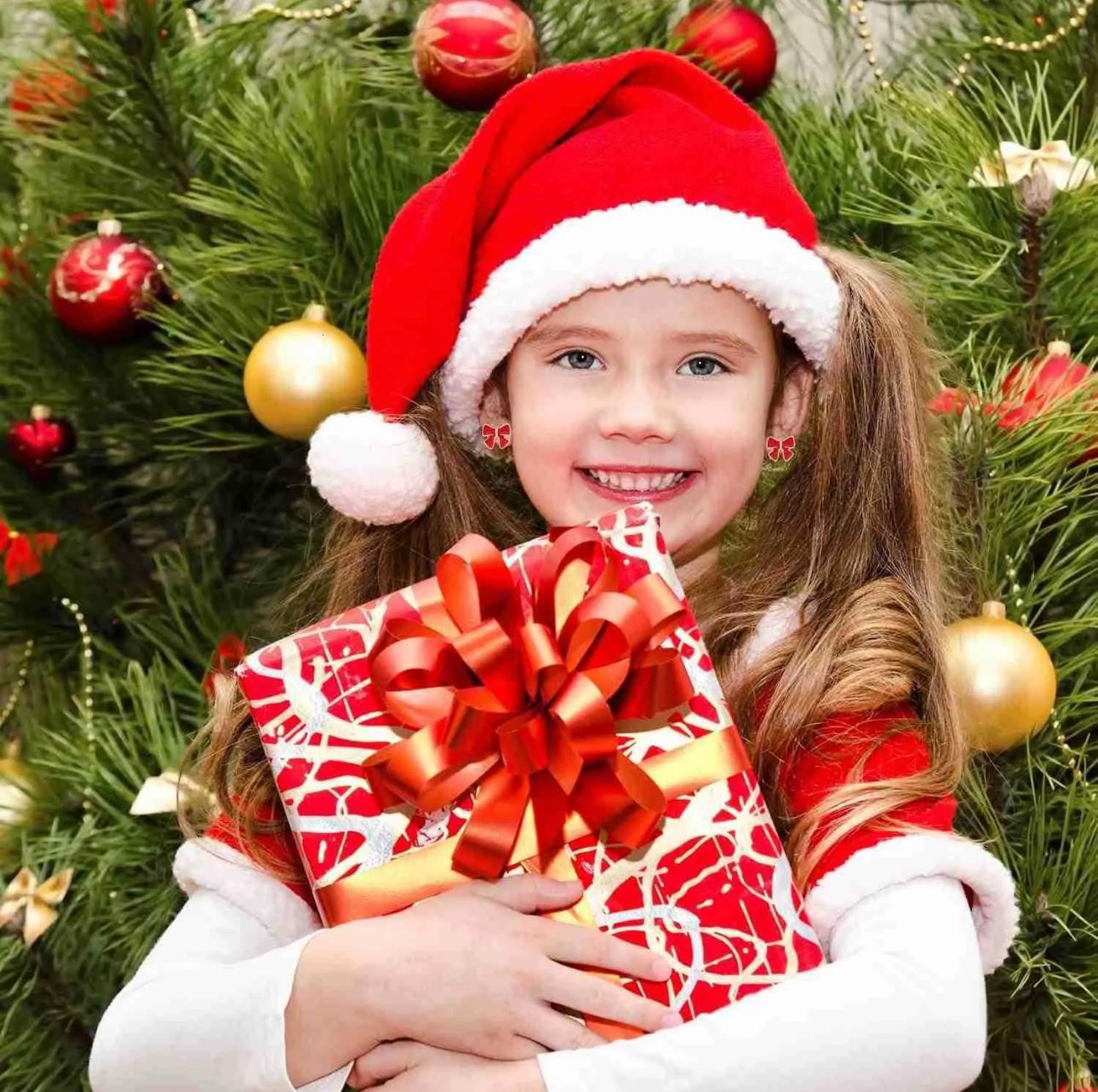 Child wearing a Santa hat holding a Christmas gift in front of a decorated tree.