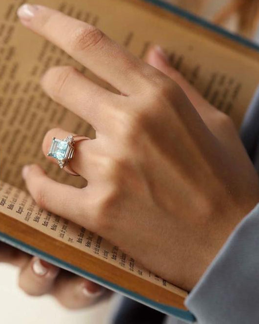 A woman's hand gracefully holding a vintage book, modeling the Aerion Celestia solid silver engagement ring featuring a massive 5.44ct emerald-cut natural sky blue topaz.