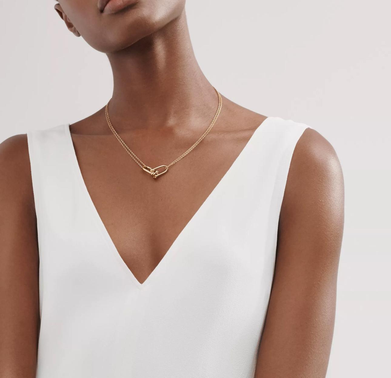 A woman wearing a gold knot pendant necklace, styled with a white V-neck dress in a minimal, elegant setting.