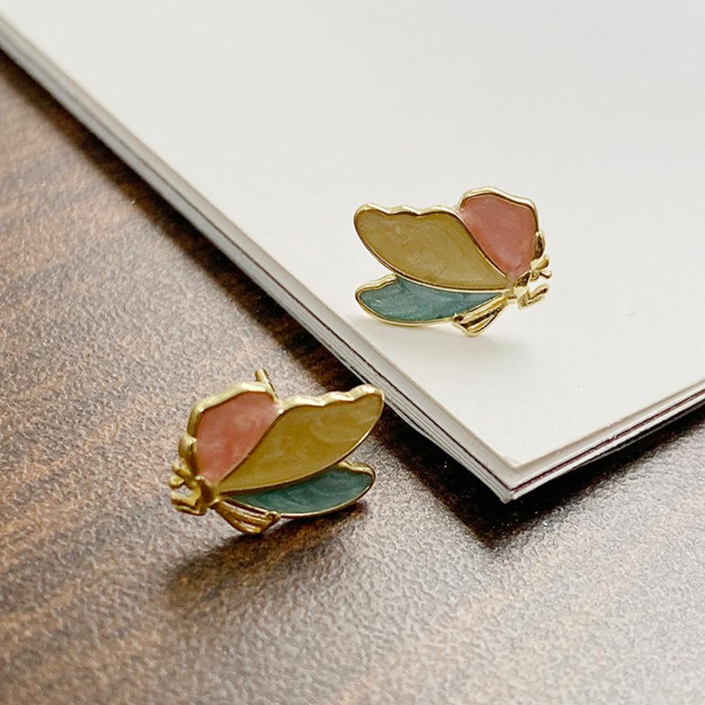Butterfly-shaped earrings with gold, green, and pink colors on a textured surface.