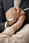 Close-up of a romantic couple holding hands, showcasing the Argénteo matching "His & Hers" solid 925 silver cuff bangles. The man wears a 12mm wide mirror-polished cuff with a tailored suit, while the woman wears a matching 8mm silver bangle with a silk blouse. Perfect luxury jewelry gift for couples.