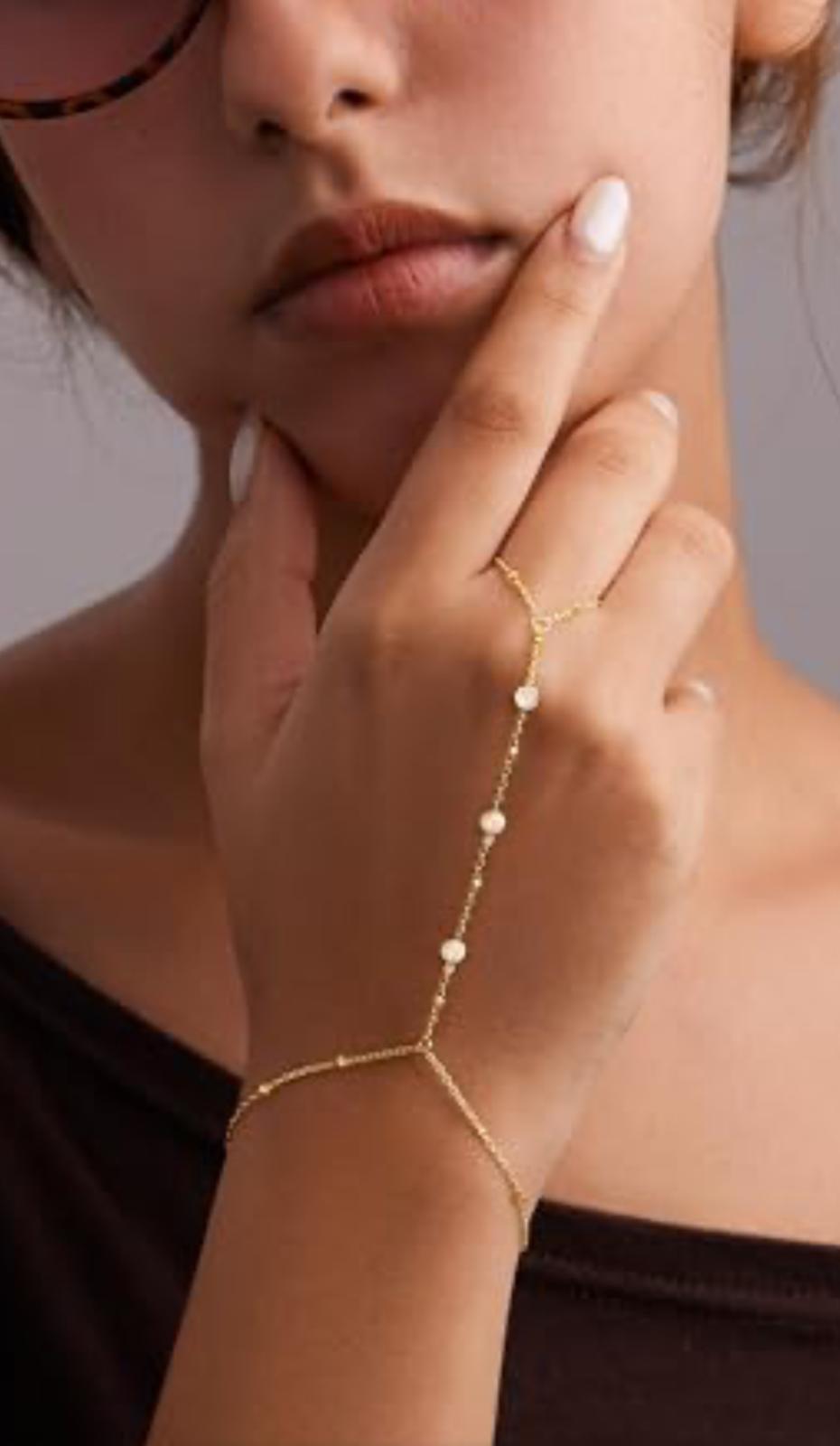 Close-up of a hand wearing a hand chain gold bracelet with small beads, touching the face.
