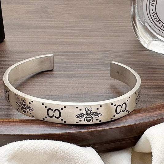 Silver bracelet with bee and 'CO' design on a wooden surface next to a perfume bottle.