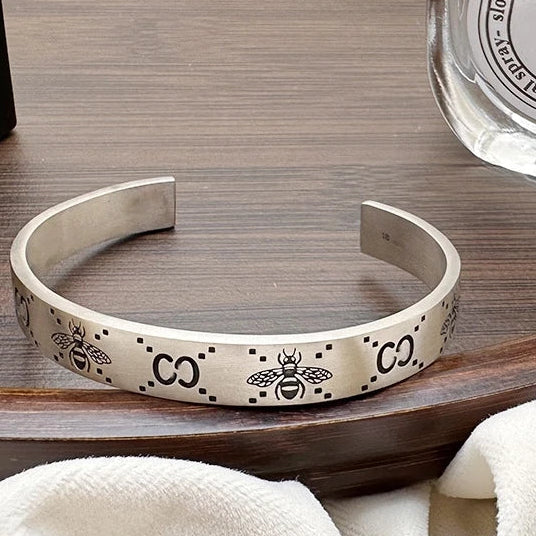 Silver bracelet with bee and 'CO' design on a wooden surface next to a perfume bottle.