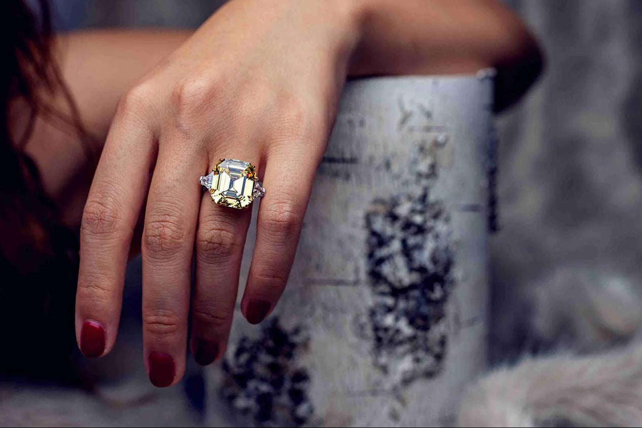 A woman's hand wearing a large emerald-cut gemstone ring with side stones, showcased in a close-up fashion shot with elegant lighting.