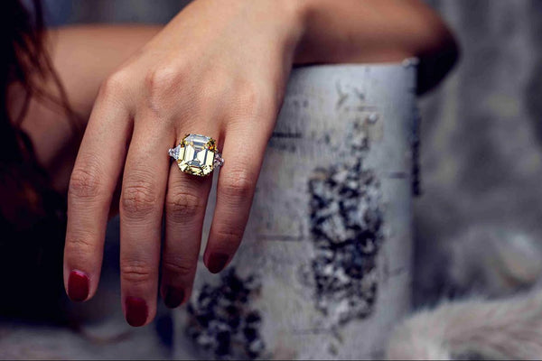 A woman's hand wearing a large emerald-cut gemstone ring with side stones, showcased in a close-up fashion shot with elegant lighting.