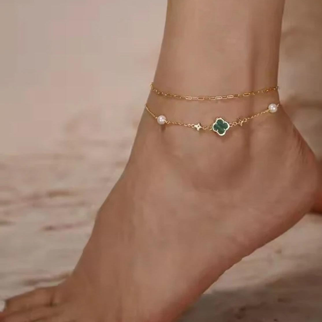 Gold anklets with green clover and pearl charms on a foot against a neutral background